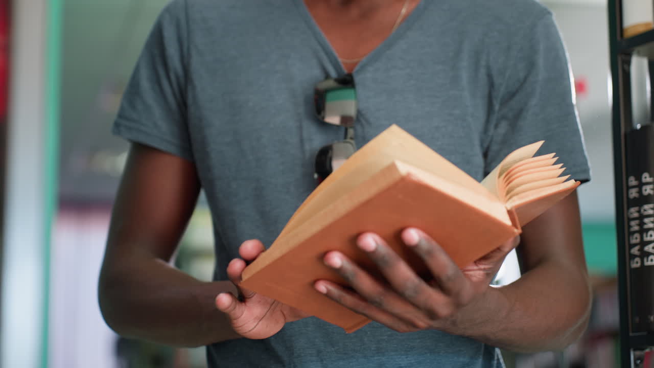 Nærbillede af mand med bagvendt kasket, der smiler, mens han læser en bog i et bibliotek, hvor sollyset blidt oplyser baggrunden og fanger et øjeblik af fokus og stille fornøjelse