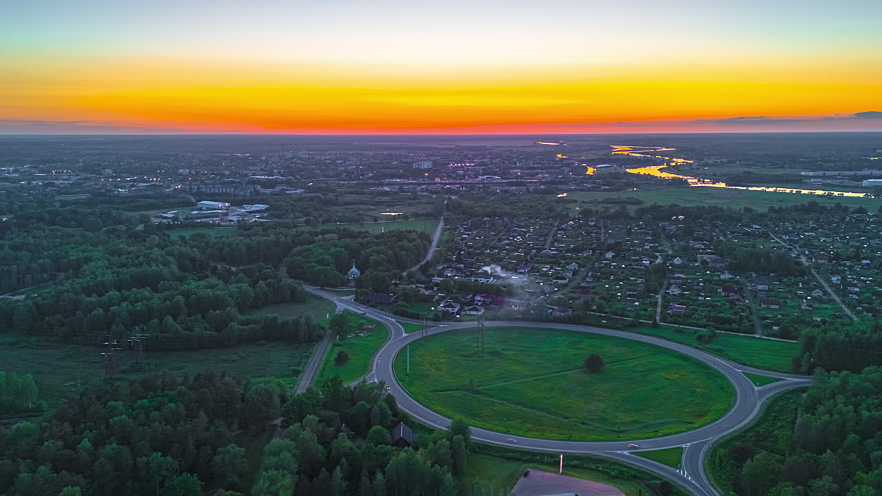 Drone hyperlapse of glowing sunset light over roundabout and rooftops in evening cityscape