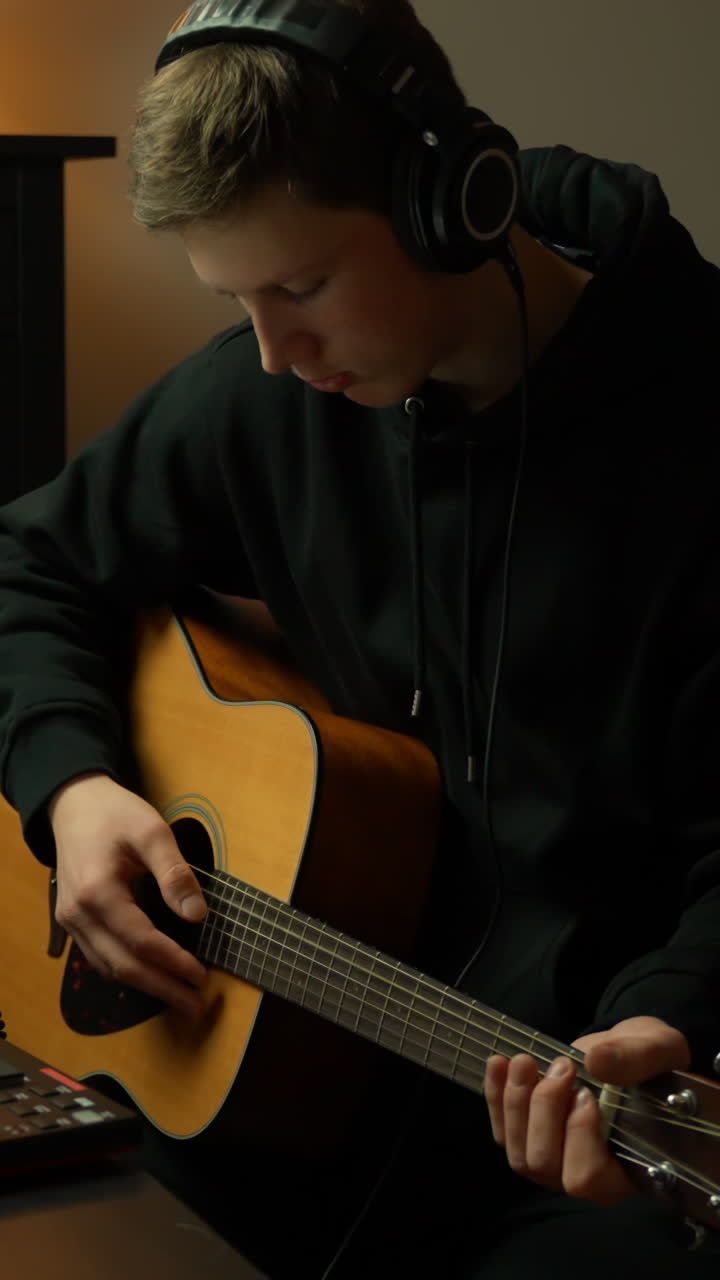 Music producer playing ascoustic guitar while composing track in dark home studio setting, vertical wearing headphones
