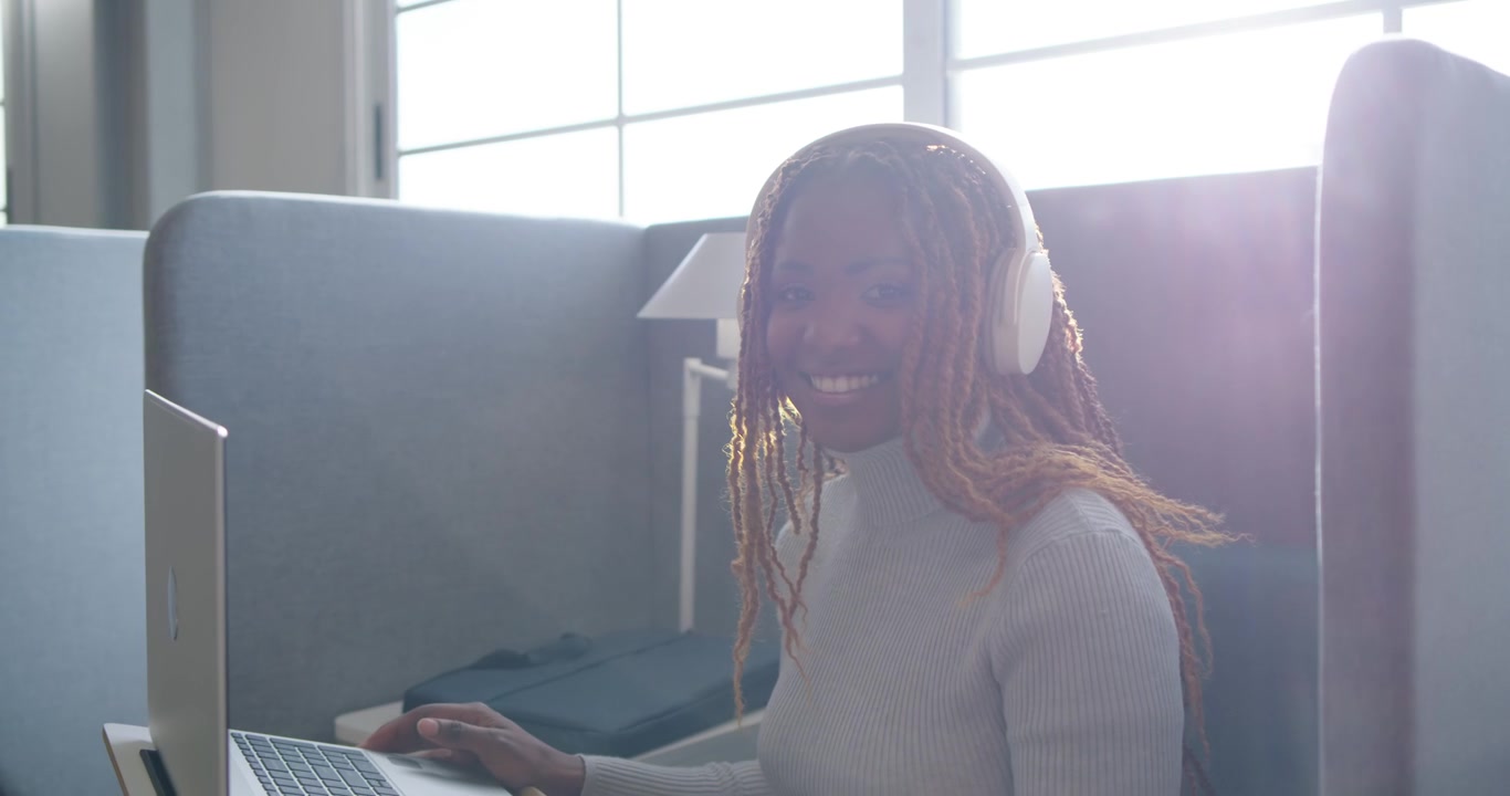 Afrikanische Frau richtet sich in Arbeitskabine ein, trägt weiße Kopfhörer, tippt auf Laptop und arbeitet remote.