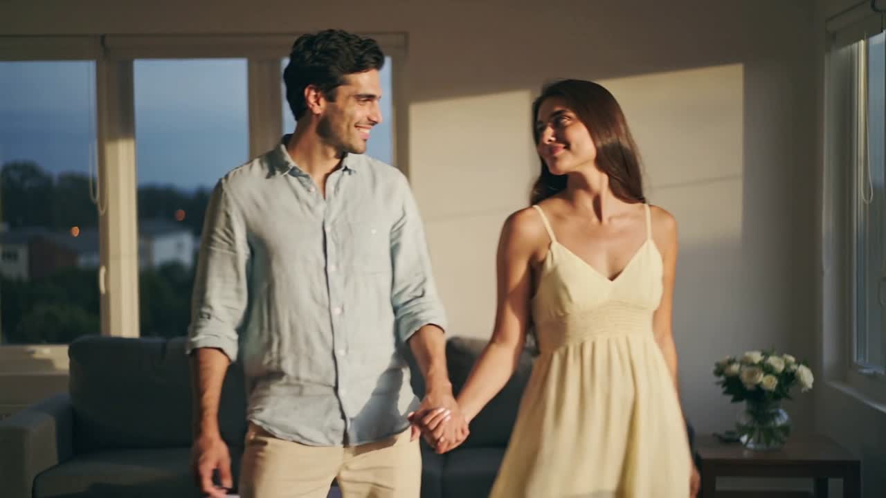Una coppia che cammina mano nella mano verso l'obiettivo a casa, scambiandosi sorrisi vicino alle finestre, lei con un vestito giallo.