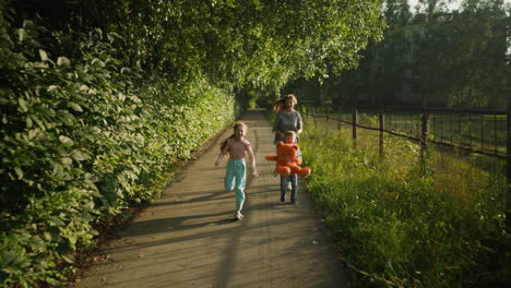 cheerful woman running with kids on paved outdoor path lined with trees and tall grass, boy holding teddy bear, girl smiling and sprinting beside them under soft morning sunlight in peaceful setting