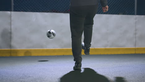 lower angle view of man kicking soccer ball against wall under bright stadium lighting at night, with urban cityscape, goal post, and illuminated buildings in the background