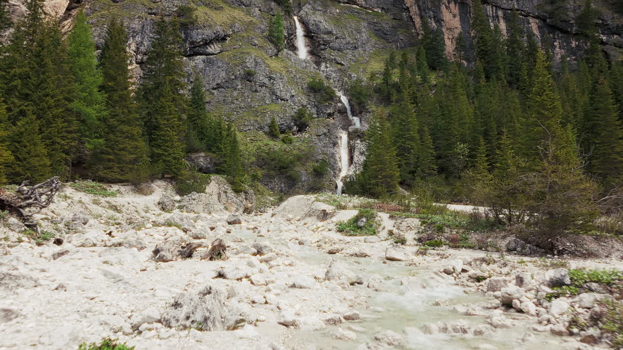 un arroyo de montaña fluye a través de un terreno rocoso con una cascada en el fondo, en los dolomitas