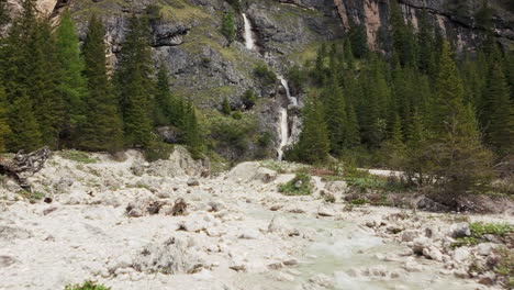 A-mountain-stream-flows-through-rocky-terrain-with-a-cascading-waterfall-in-the-background,-set-in-the-Dolomites