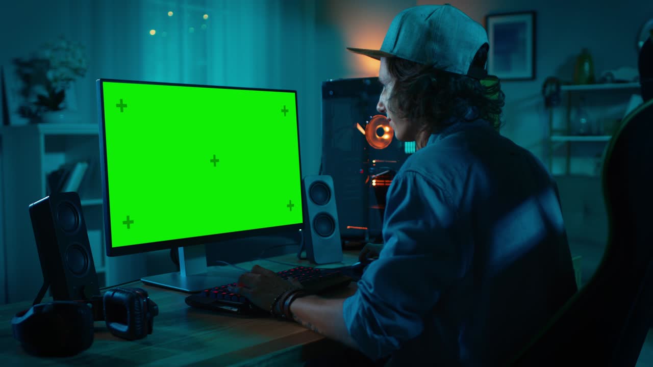 Gamer Profesional bermain video game online di komputer pribadinya yang canggih dengan lampu LED neon berwarna-warni. Layar Hijau Mock-up. Pria muda mengenakan topi. Ruang tamu dengan lampu hangat. Malam hari