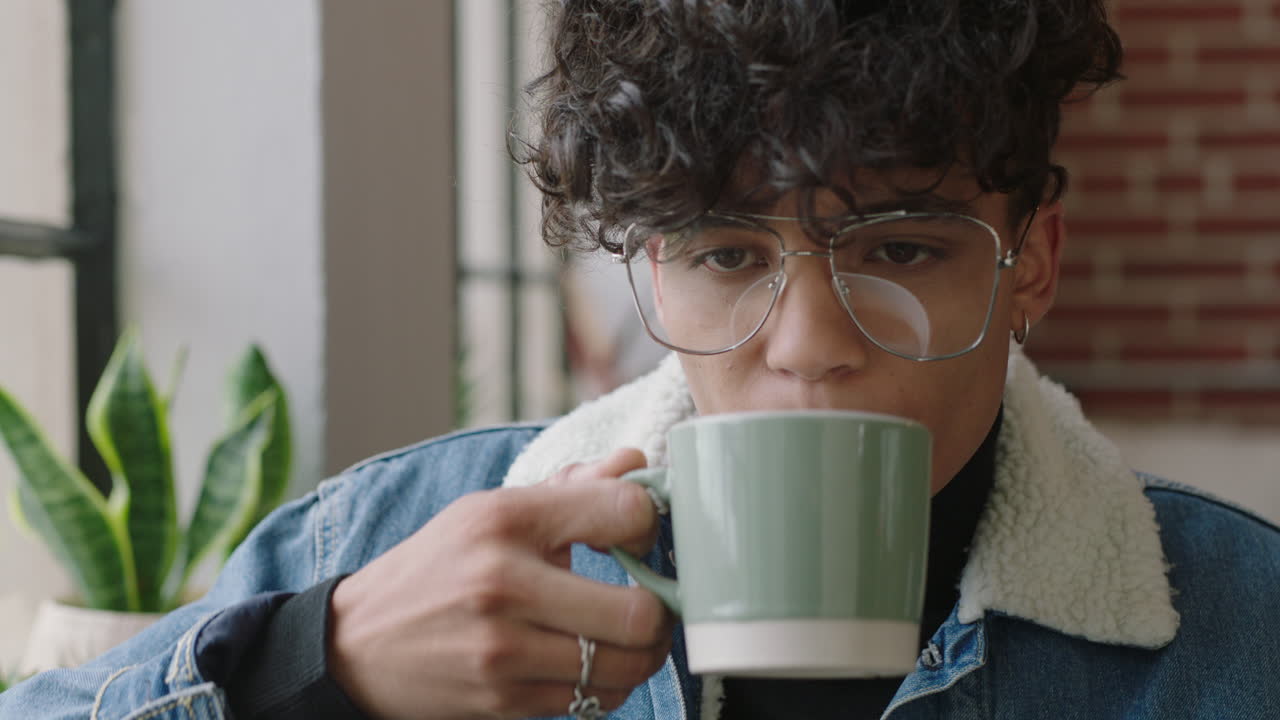 joven hispano con estilo estudiante bebiendo café en casa mirando por la ventana pensando en el futuro disfrutando de un estilo de vida relajado usando gafas de moda de moda