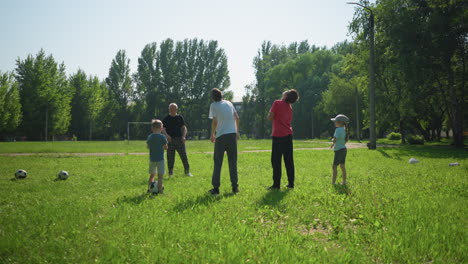 a grandfather is leading his two grandsons in a stretching exercise on a grassy field, one grandson holds a ball between his legs while they all turn their necks as the later jump together