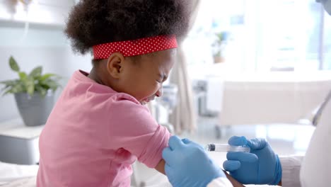 african american female doctor with face mask making injection to girl in hospital room, slow motion