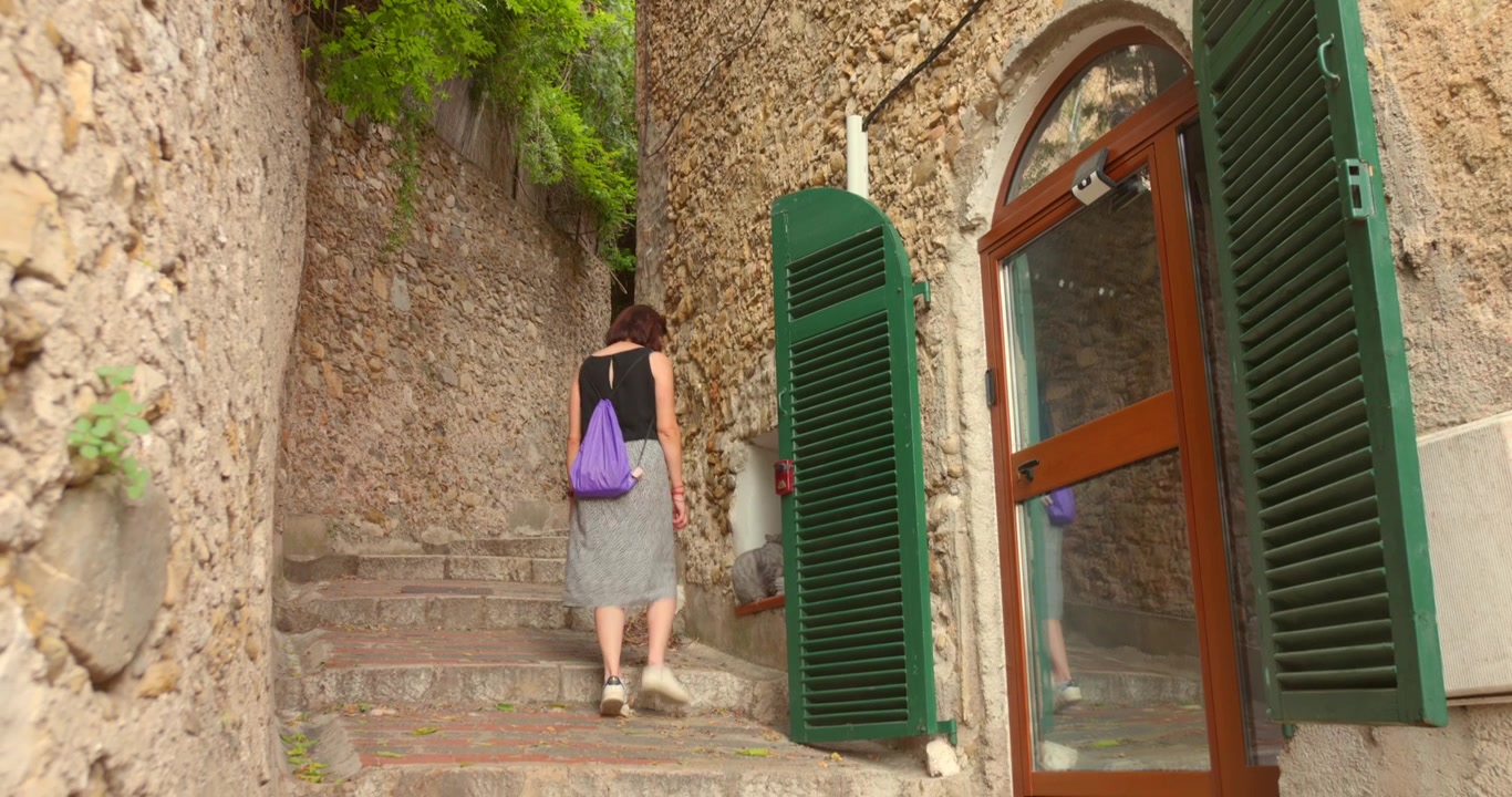 Donna che sale gradini di pietra tra vecchie mura di un villaggio medievale accanto a persiane verdi nell'incantevole Roquebrune-Cap-Martin sulla Costa Azzurra, Francia