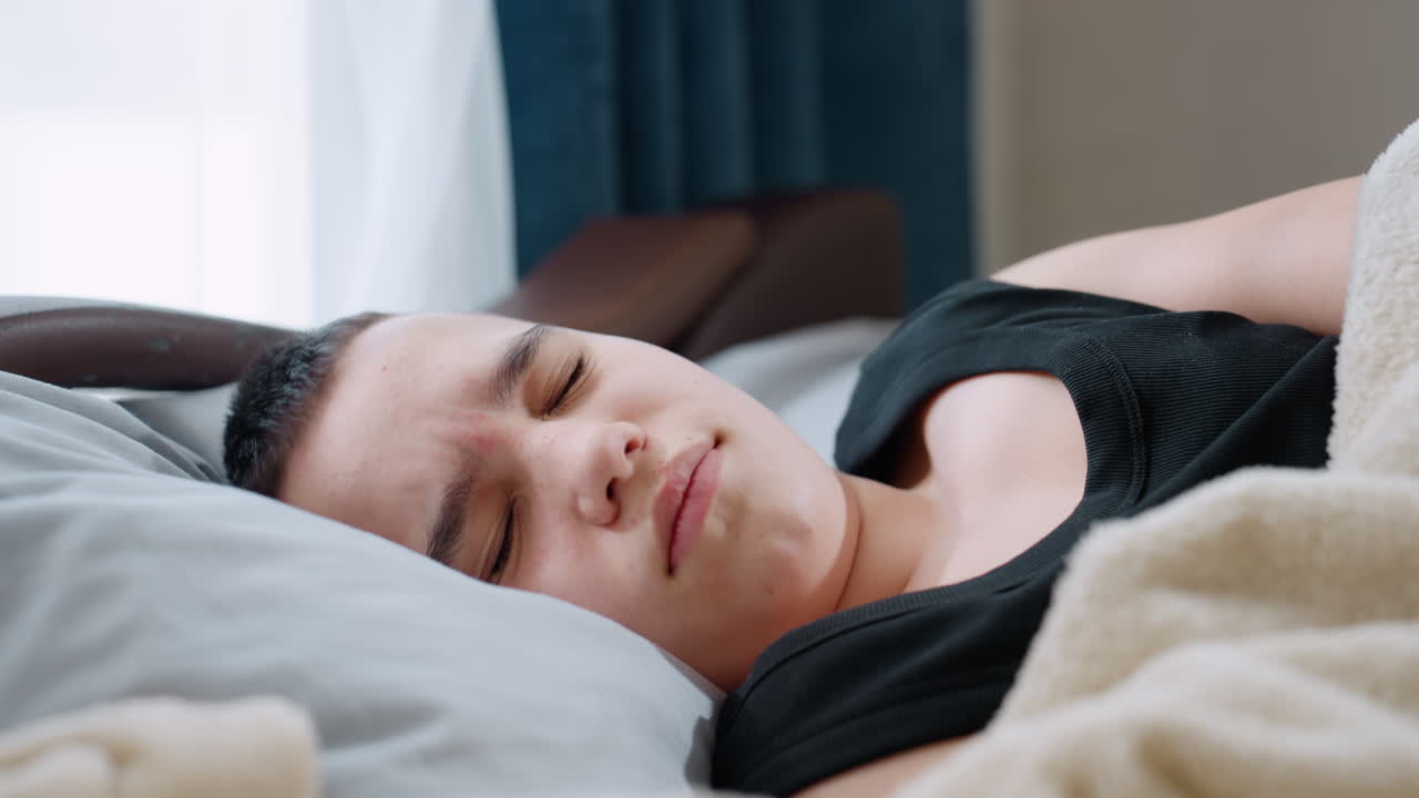Close up boy in black singlet sleeping on pillow looking restless with closed eyes showing discomfort and uneasy rest under blanket in bedroom highlighting tired child