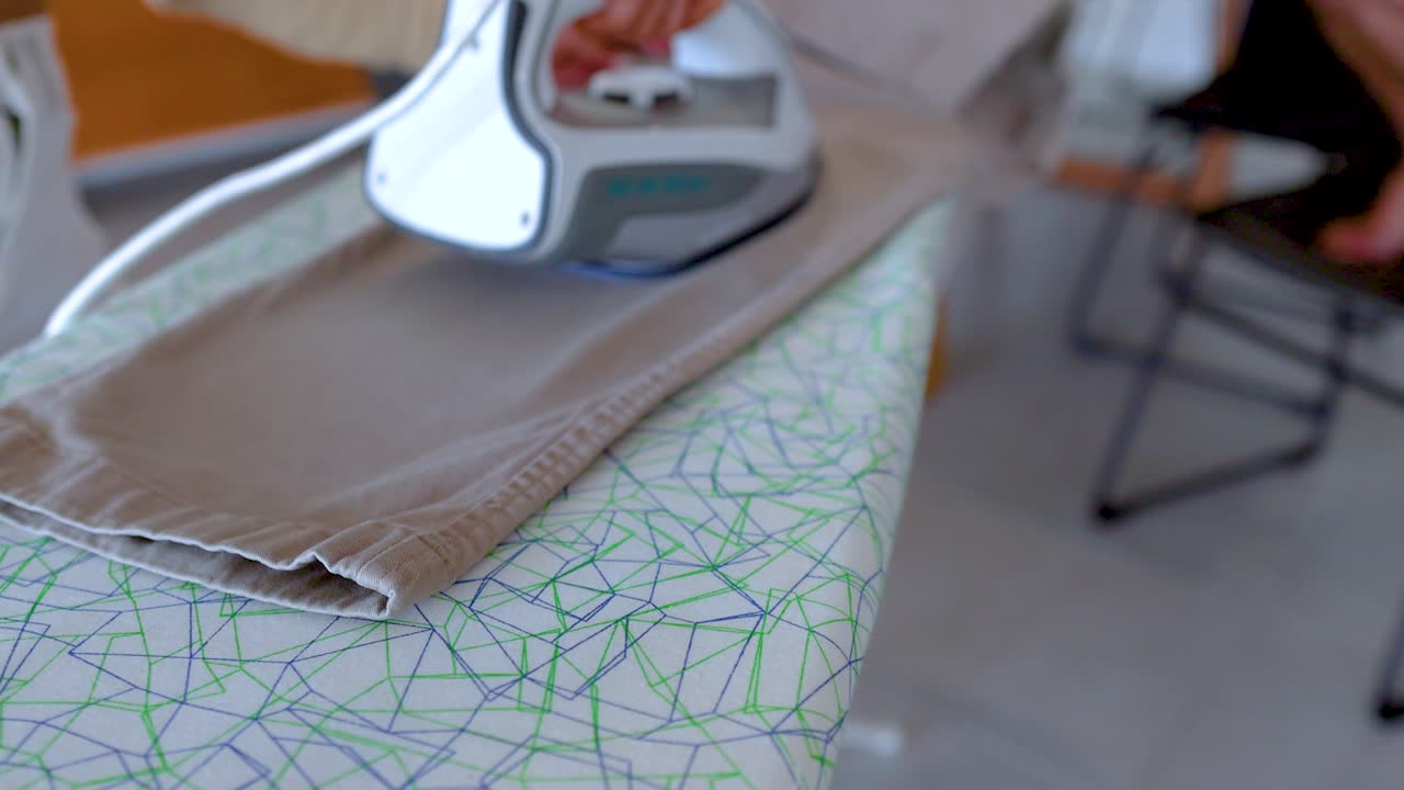 A person irons beige pants on a patterned board in a well-lit room, focusing on smooth fabric