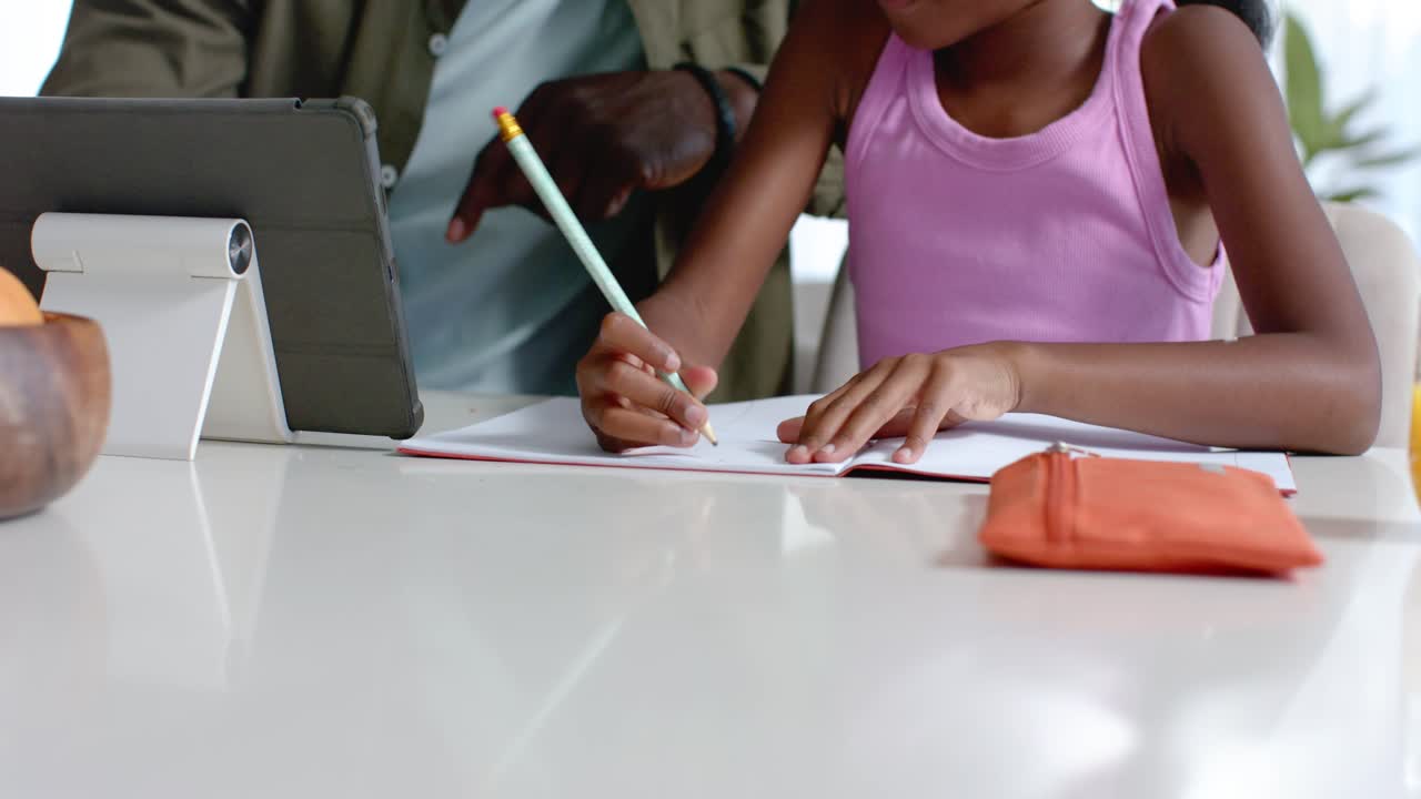 Mature guardian pointing at tablet, guiding youth female writing with pencil in notebook at table
