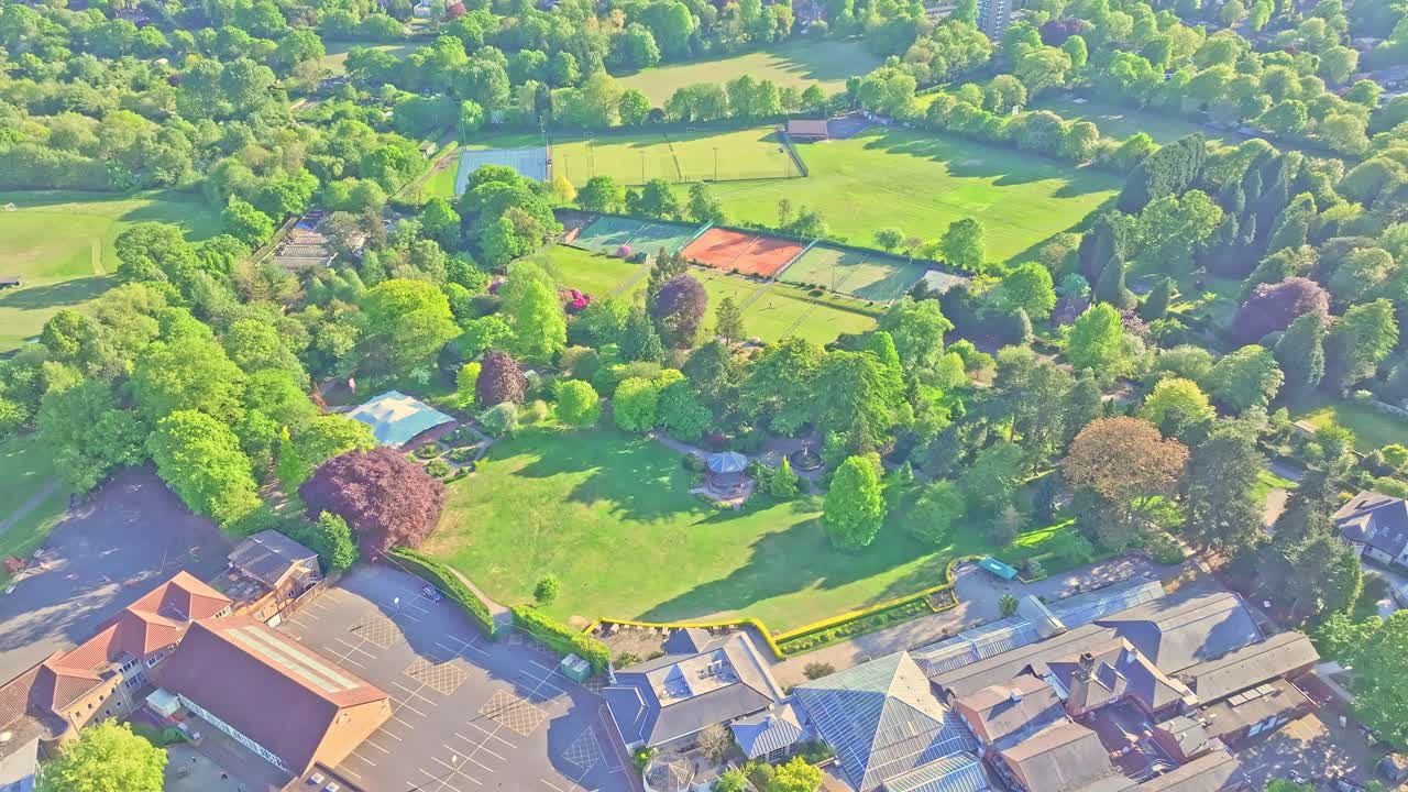 Drone slow push-in over Birmingham Botanical Gardens, featuring the bandstand, glasshouses, lawns, tennis courts, and surrounding greenery in Edgbaston