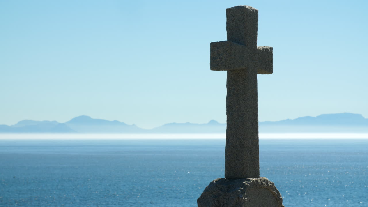 Tombstone memorial in right of frame overlooking ocean with layer of mist and blue mountains in distant horizon on clear sunny day