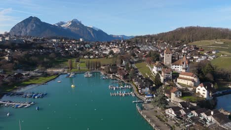 Ciudad-De-Spiez-Con-El-Lago-Thun-Y-Los-Alpes-Suizos-Al-Fondo,-Cielo-Despejado,-Barcos-En-El-Agua,-Vista-Aérea