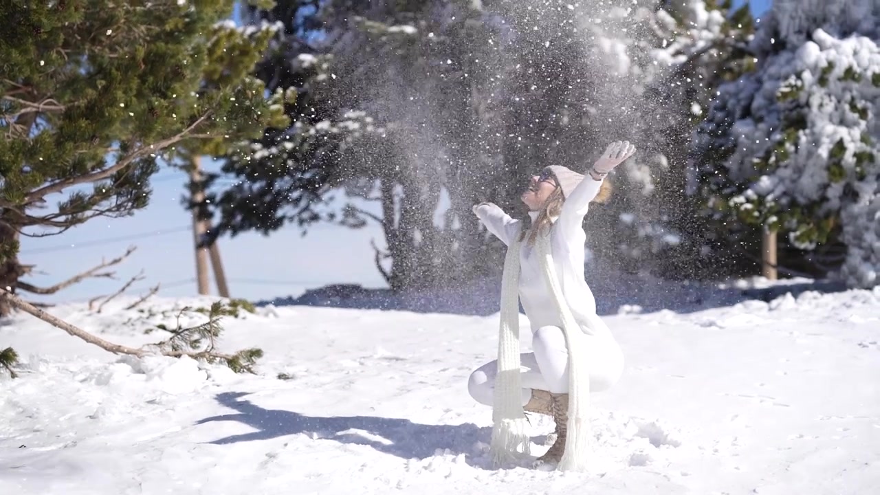 Eine positive Frau mittleren Alters in warmer Kleidung wirft im Winter Schnee in einem verschneiten Wald in die Luft.
