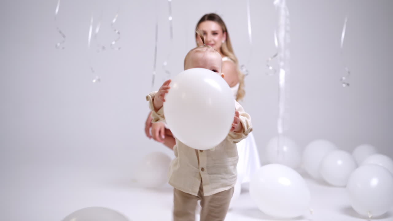 Adorable happy baby walks unsteadily holding a balloon. Smiling mother sits behind watching her beloved son. Studio footage.