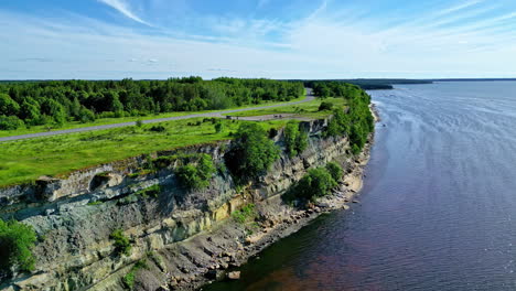 Breathtaking-aerial-view-of-Türisalu-Cliff,-Estonia,-on-a-sunny-summer-day-overlooking-the-calm-body-of-water,-with-lush-green-trees-and-foliage-in-the-foreground