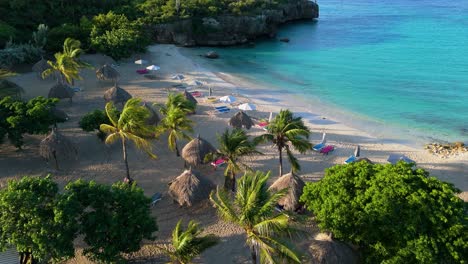 Aerial-dolly-above-tropical-beach-side-huts-and-palm-trees-with-long-shadows,-ocean-waves-crash-on-Daaibooi-beach