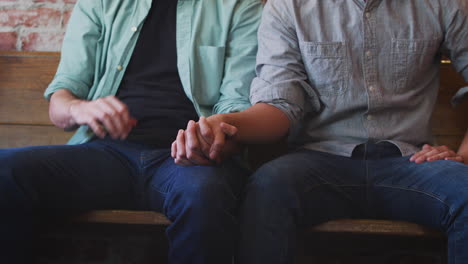 close up of same sex male couple holding hands sitting on bench in coffee shop together