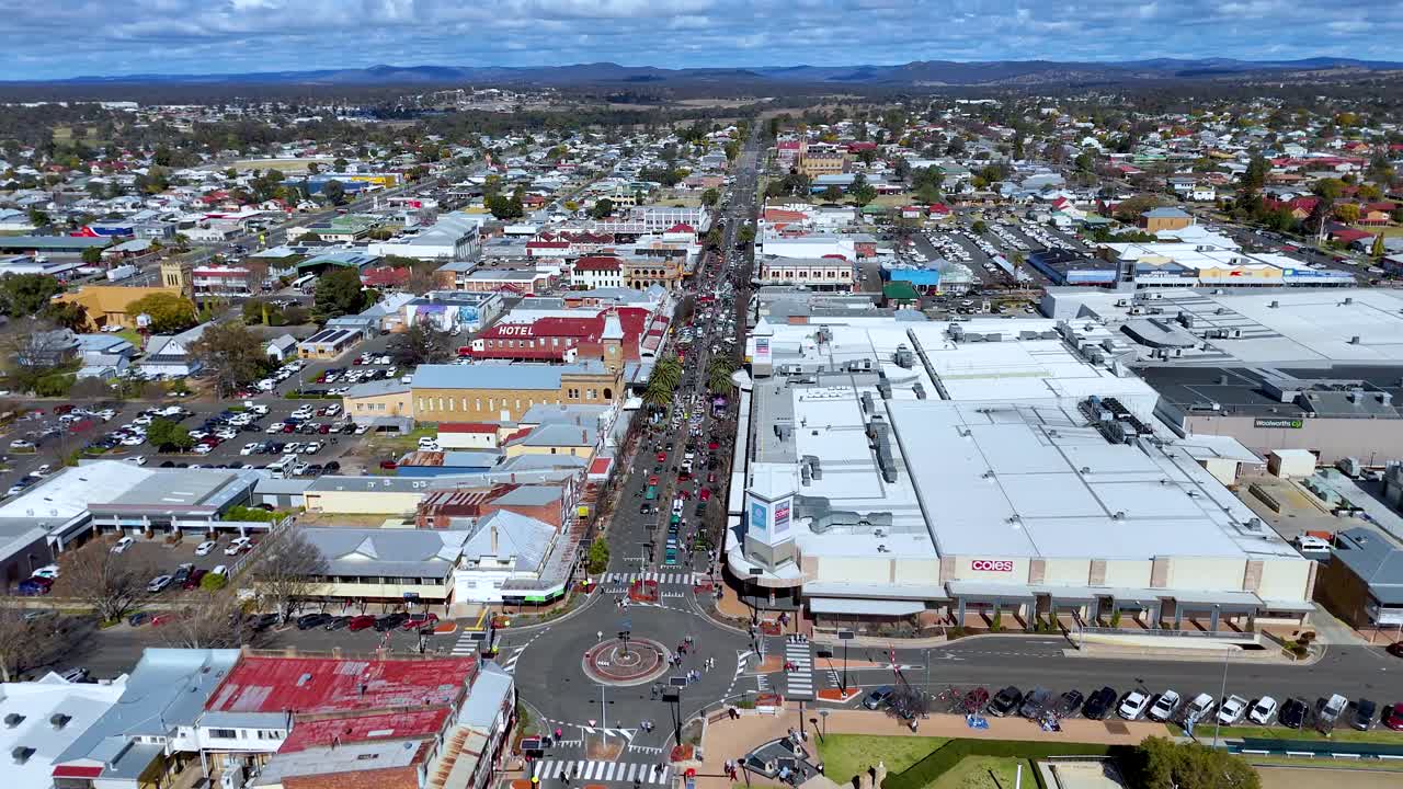 Drone footage glides above a vibrant town center, capturing a roundabout, festival crowds, parked cars, and colorful buildings under bright daylight