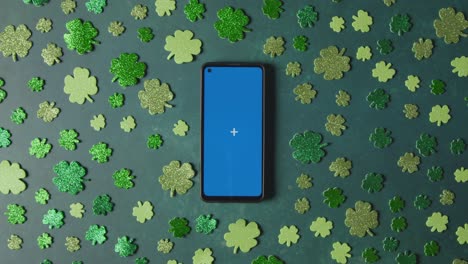 foto de estudio superior de formas de trébol verde que rodean el teléfono móvil de pantalla azul para celebrar el día de san patricio