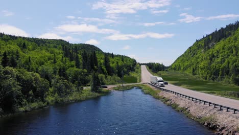 A-white-semi-truck-drives-down-a-highway-surrounded-by-forests-and-mountains-next-to-a-lake-during-the-day-in-Ontario,-Canada