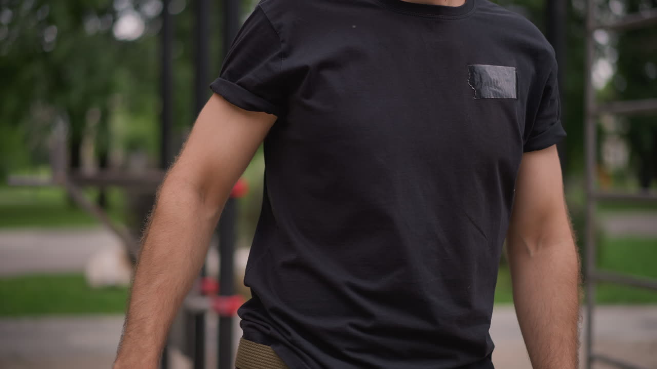 Man Attaching Red Resistance Band To Bar In Outdoor Park, Focused Urban Trainer Secures Loop On Metal Pull Up Rig, Closeup Grip And Forearm Tension, Leafy Background, Midday Light, Quiet Dedication