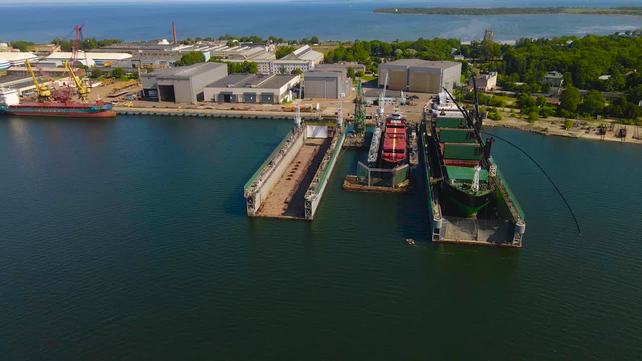 Aerial drone footage orbiting around a large and sunny summer shipyard where a massive cargo or container ship boat is repaired and being built. Ship is raised and separated from blue ocean water