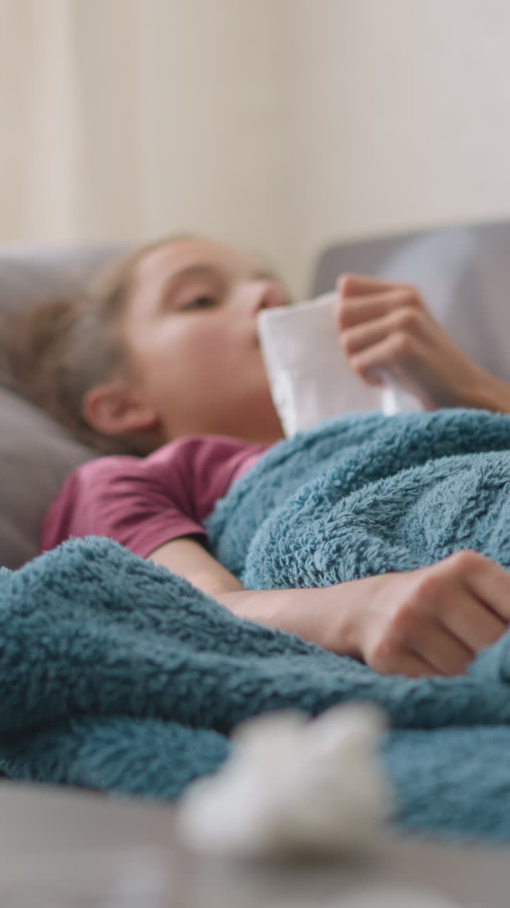 Primo piano di una ragazza malata che starnutisce in un fazzoletto mentre è sdraiata sul divano sotto una coperta calda con una tazza di vetro e fazzoletti usati sul tavolo in primo piano, a indicare influenza, raffreddore o guarigione da una malattia stagionale