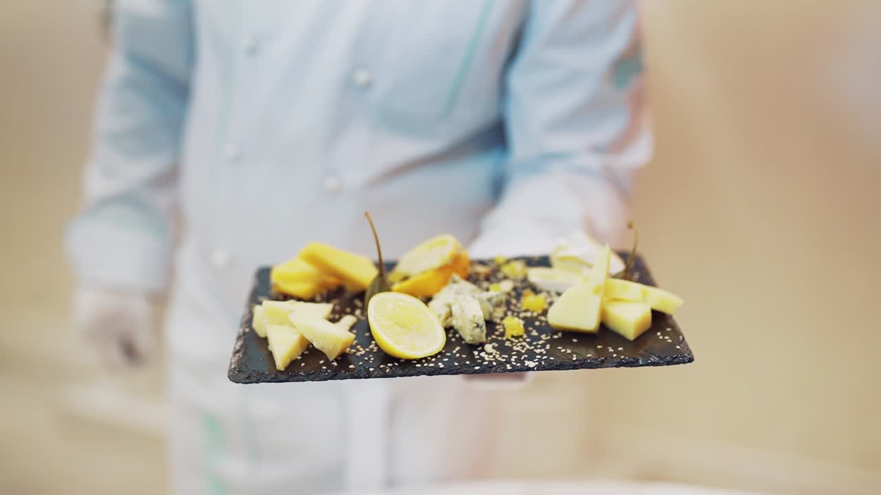 Professional chef in white clothes decorates the dish with lemon on the black board. Different kinds of tasty cheese nicely presented by cook in the restaurant.