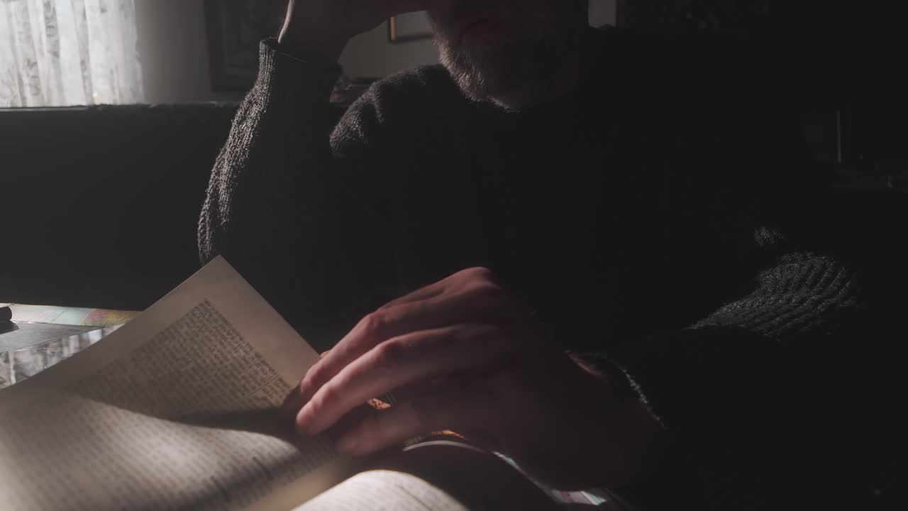 Nonchalant bearded man in dimly lit shadows idly turning book pages Medium shot