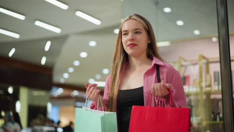 beautiful lady in pink dress lifts her red and mint green shopping bags with a contented smile, taking a deep breath, she stands in a brightly lit shopping mall, with other shoppers in the background