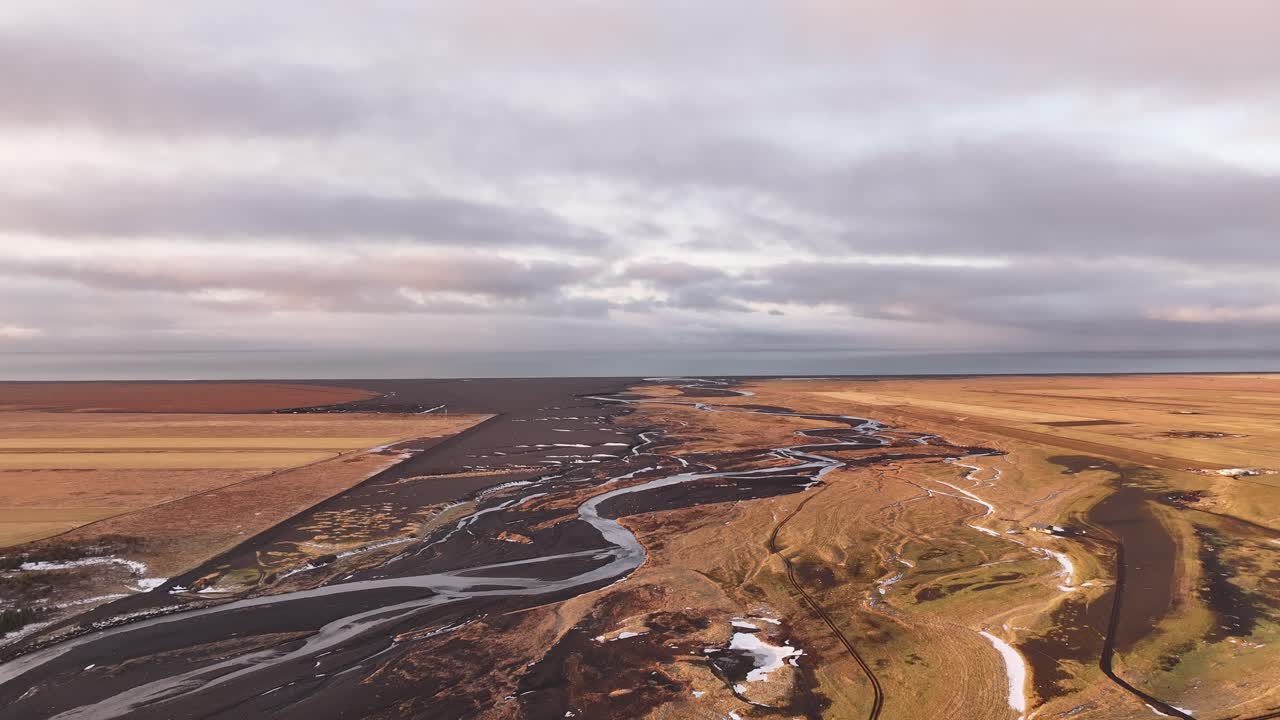 Drone advances over braided glacial rivers flowing toward Iceland's southern coast near Skógar, revealing vast golden plains under a dramatic cloudy sky.
