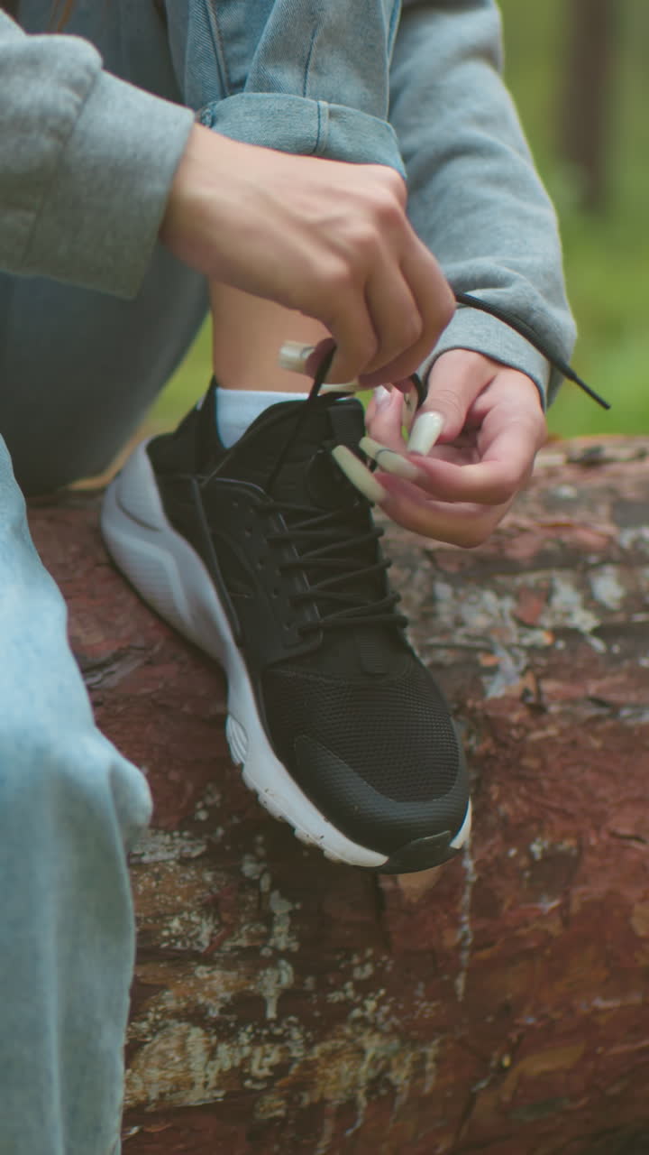 긴 손을 가진 여성이 산책하는 동안 쓰러진 나무에 앉아 신발을 는 클로즈업, 그녀는 회색 스웨터와 밝은 파란색 청바지를 입고, 숲에서 검은 운동화를 조정하는 데 집중합니다.