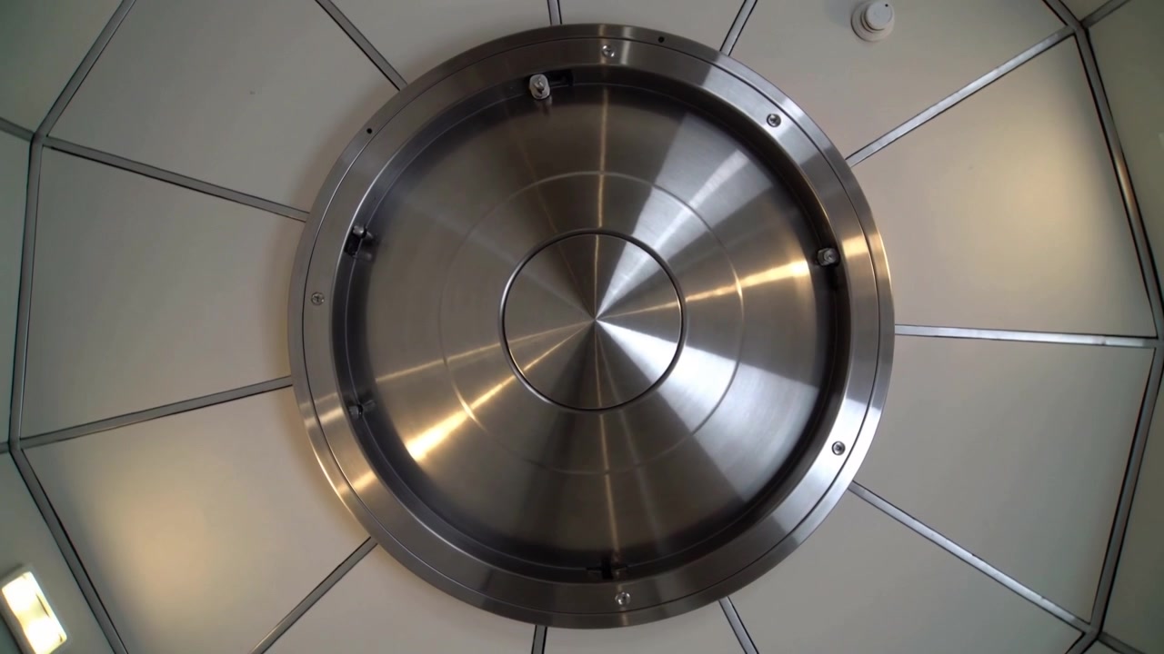 Panel de techo metálico circular giratorio moviéndose en el sentido de las agujas del reloj en una sala abovedada, mostrando un sensor y una luz.
