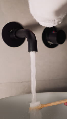mujer lava el cepillo de dientes con agua corriente en el baño de primer plano. dama en bata de baño de terry suave limpia el cepillo después de lavarse los dientes en casa. higiene personal