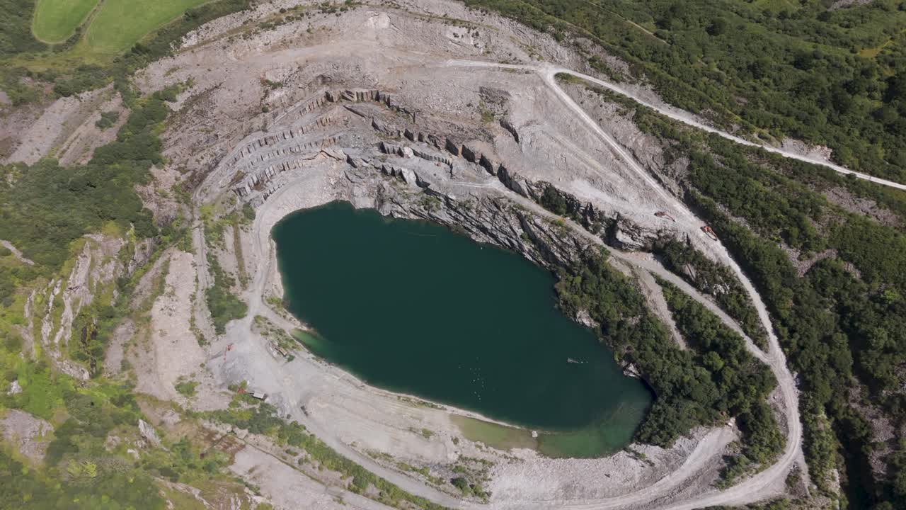 Aerial view of a remote flooded slate quarry, featuring steep terraced rock formations, calm green water, and natural vegetation, Cornwall, UK