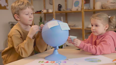 blond boy and blonde little girl in craft workshop sticking notes with environmental quotes on a blue globe