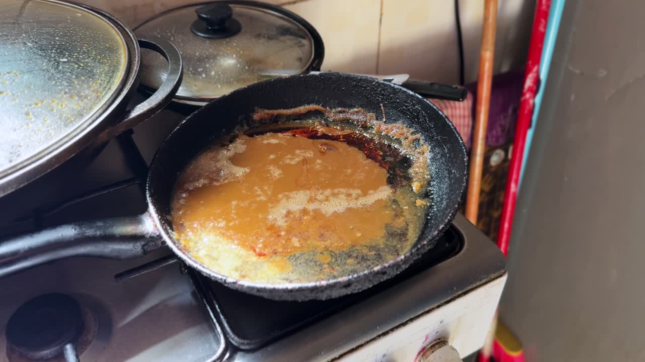 Close-up of a frying pan filled with burnt cooking oil and residue on a home gas stove, with covered pots and stained cookware nearby, showing greasy cooking mess and cleanup needs