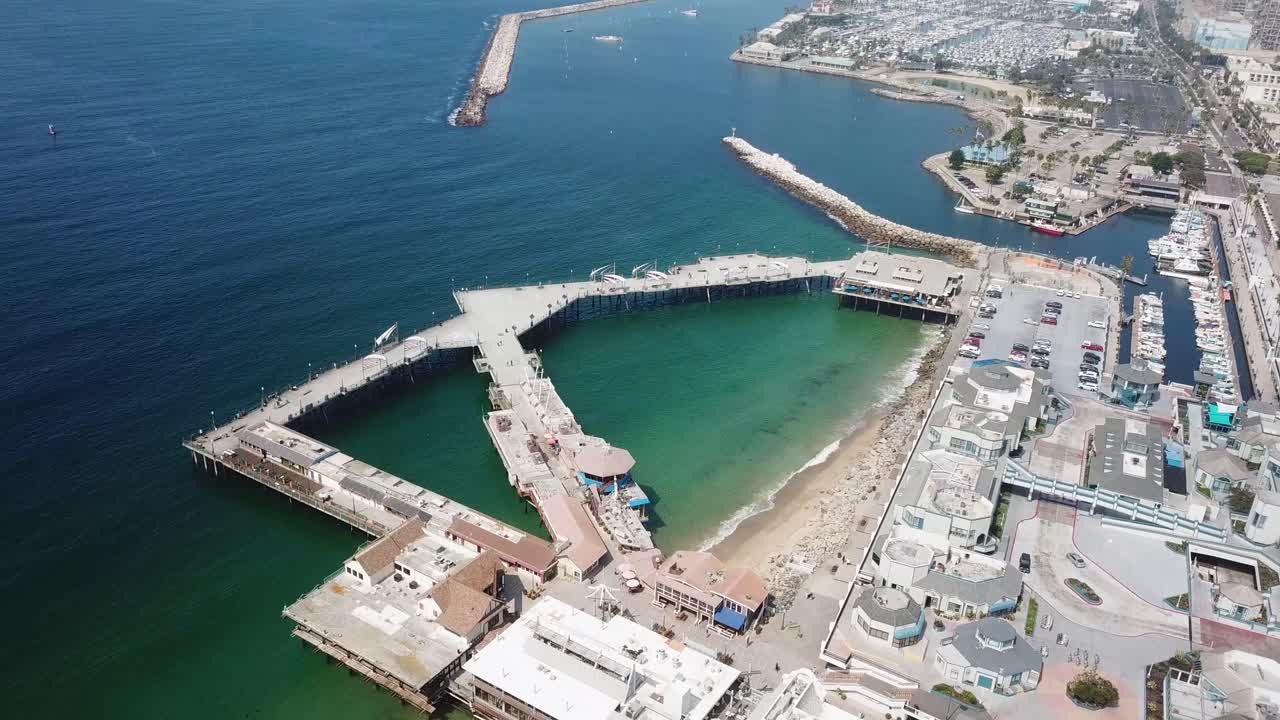 Drone pushes in over the horseshoe pier at Redondo Beach revealing the protected cove breakwater and adjacent harbor on a bright late August day. Clear green water sandy shoreline and waterfront shops