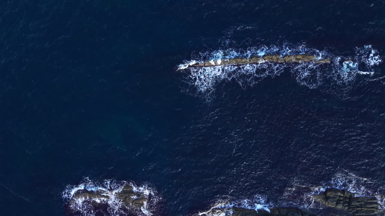 Waves dance around rocky formations under a vast blue sky at the seashore, capturing the beauty of nature's coastal wonders