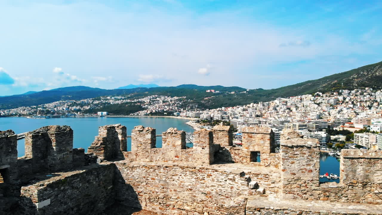 Aerial view of Kavala, a lot of buildings, Aegean sea coast, green hills in the distance. Drone flies out from behind the wall of ancient fort. Greece