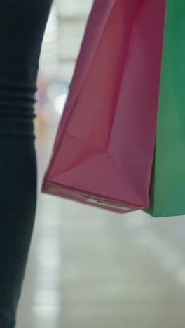 vista de cerca de la pierna de una mujer en vaqueros y zapatillas de lona que lleva una bolsa de compras de menta y rosa en su mano derecha mientras camina en un centro comercial bien iluminado