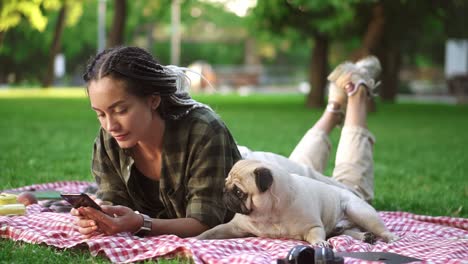 Mujer-Acostada-Al-Aire-Libre-En-El-Picnic-Plaid-En-El-Césped-Usando-Su-Teléfono-Inteligente-Y-Un-Pequeño-Y-Lindo-Pug-Está-Acostado-Cerca