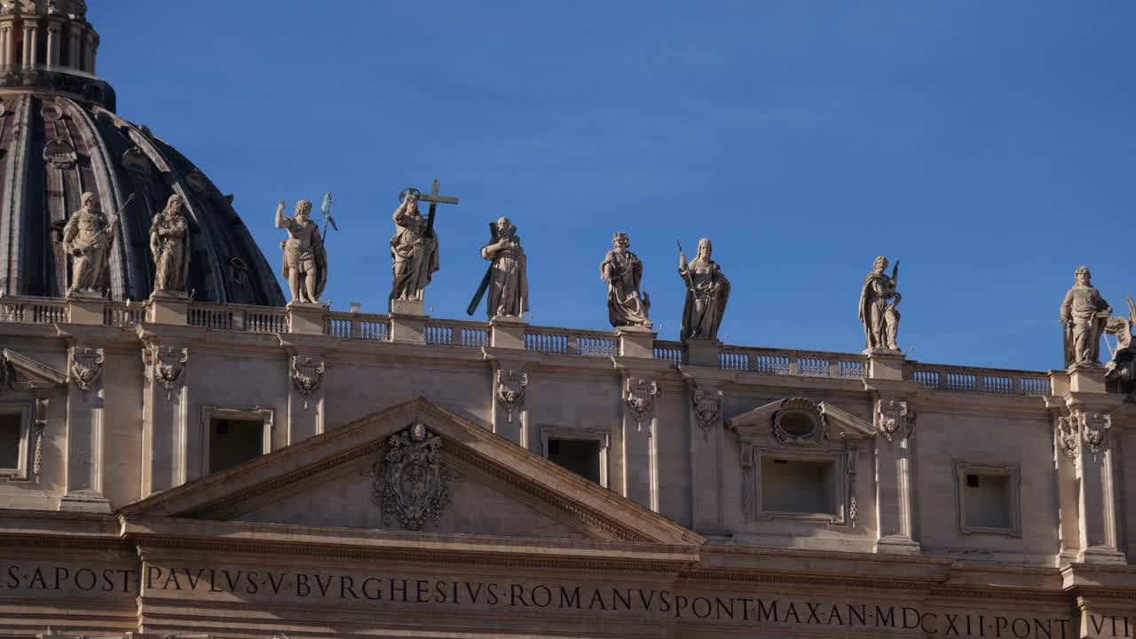 Statues of apostles, including figures with a cross, line the roof above St. Peter’s Basilica’s entrance in Vatican City, Rome, captured in bright sunlight and clear blue skies