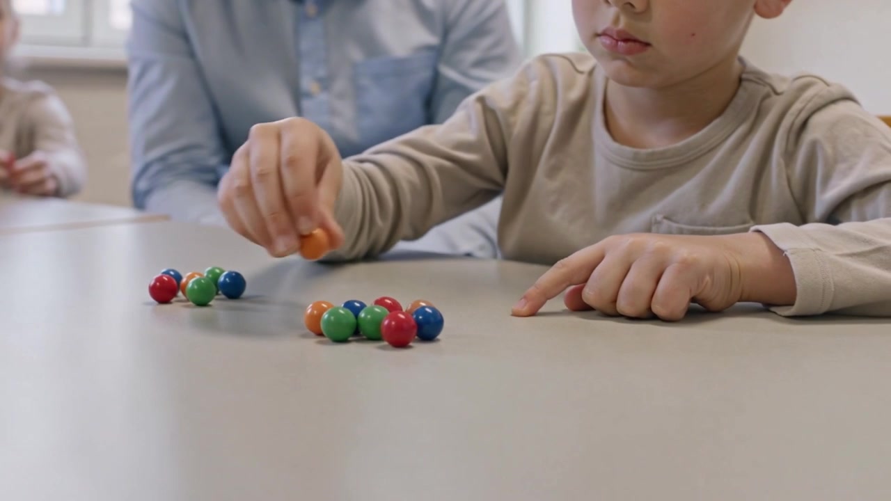 Bambino con maglietta beige che indica e raccoglie una perlina al tavolo della scuola per contare mentre l'insegnante osserva, spazio per il testo
