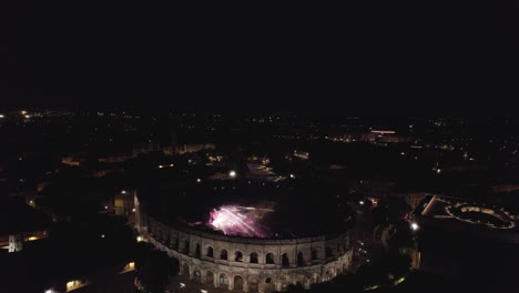 Rotación-De-Drones-En-Las-Arenas-De-Nîmes-En-Medio-De-La-Noche,-La-Gente-Está-Viendo-El-Concierto-Y-Hay-Luces-De-Varios-Colores