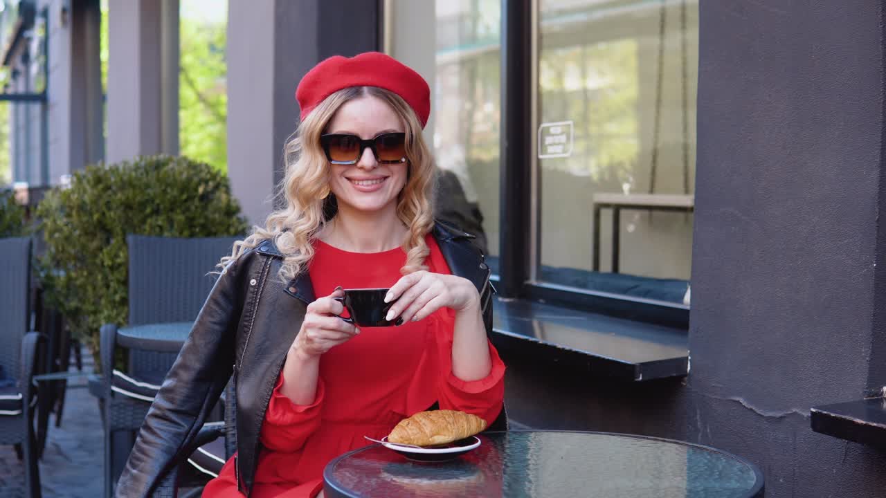 Stylish coffee. The blonde drinks coffee with a croissant. Modern black terrace of the restaurant. Young woman in sunglasses and beret
